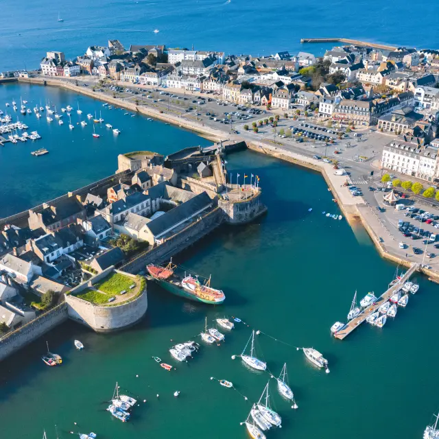 Une photo aérienne d’un port. La photo montre une ville côtière avec un bassin portuaire rempli de voiliers, des quais, un parking et des bâtiments serrés le long du front de mer; au premier plan, une zone fortifiée/enceinte en pierre au bord de l’eau et plusieurs bateaux au mouillage.