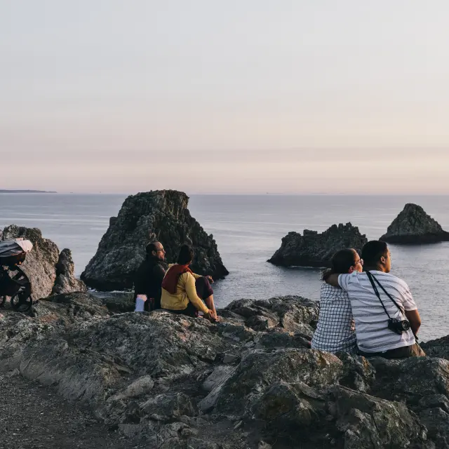 Une grande photo de bord de mer au coucher du soleil. Des rochers au premier plan, la mer calme au milieu, et plusieurs grands rochers/îlots au large. Sur les rochers, plusieurs personnes assises de dos, et une poussette est posée à gauche.