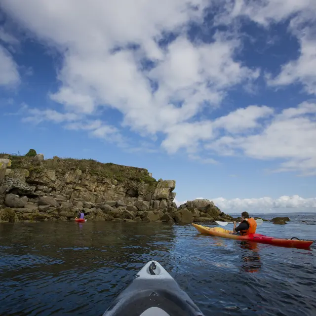 Une photo de mer à gauche qui montre un plan d’eau calme avec des kayaks, un rocher à gauche, et un ciel bleu avec de nombreux nuages blancs; la pointe d’un kayak gris apparaît au premier plan en bas.