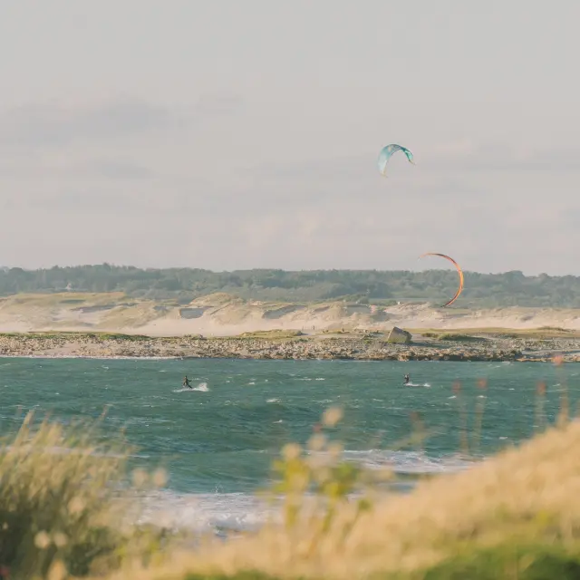 Une photo qui montre un paysage côtier: mer bleu-vert avec des vagues, une bande de dunes/sable et végétation au loin, et deux cerfs-volants de kitesurf dans le ciel (un bleu en haut, un orangé plus bas). Des herbes floues au premier plan.