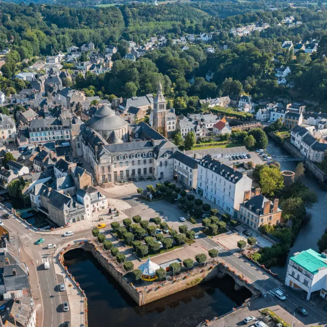 Une photo qui montre une vue aérienne d’une ville avec de nombreux bâtiments, une grande église au centre, un cours d’eau sombre au premier plan, des routes et des arbres, et un vaste paysage verdoyant jusqu’à l’horizon sous un ciel bleu.
