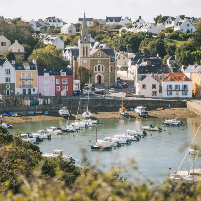 Une photo d’un port : eau calme, plusieurs bateaux (voiliers et petites embarcations) au mouillage, des maisons blanches et colorées en bord de quai, et une église au centre du village sur la colline.