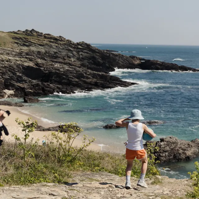 Une photo de paysage côtier. La scène montre une côte rocheuse avec une petite plage et une mer bleue; au premier plan, deux enfants sont de dos, l’un au centre avec un chapeau clair, débardeur blanc et short orange, l’autre à gauche en tenue à motifs avec bottes bleues.