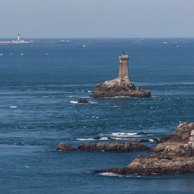 Une photo de mer avec des rochers et un phare sur un îlot (au centre), plus une balise en mer à gauche, et des rochers au premier plan à droite.