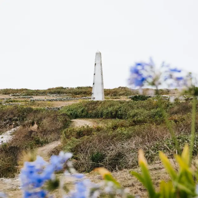 Une photo de paysage qui montre une lande côtière avec un chemin, de la végétation basse, des fleurs bleues floues au premier plan, et une haute balise/amer blanc vertical au centre-gauche sur fond de ciel clair.