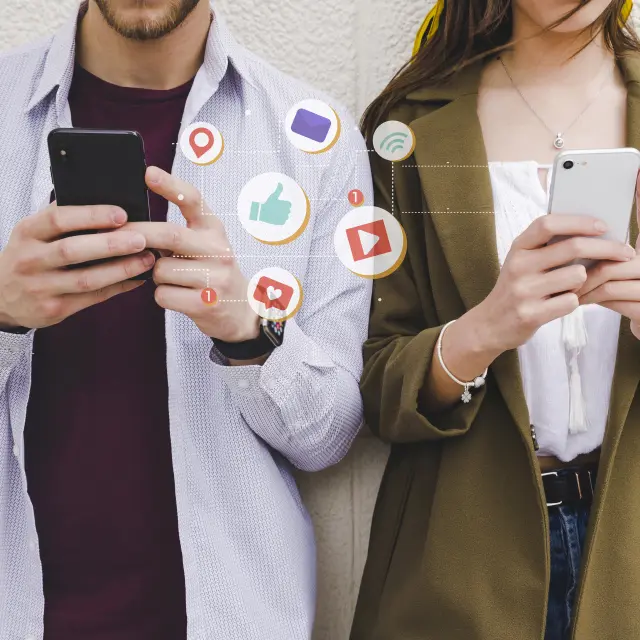 La photo montre deux adultes coupés au niveau du visage (seuls le bas du visage/menton et le torse sont visibles), debout devant un mur clair, chacun tenant un smartphone (un téléphone noir à gauche, un téléphone blanc à droite). Plusieurs icônes rondes de type “réseaux sociaux” flottent entre eux (localisation, message/bulle, symbole Wi‑Fi, pouce levé, bouton lecture/vidéo, cœur/like, etc.).
