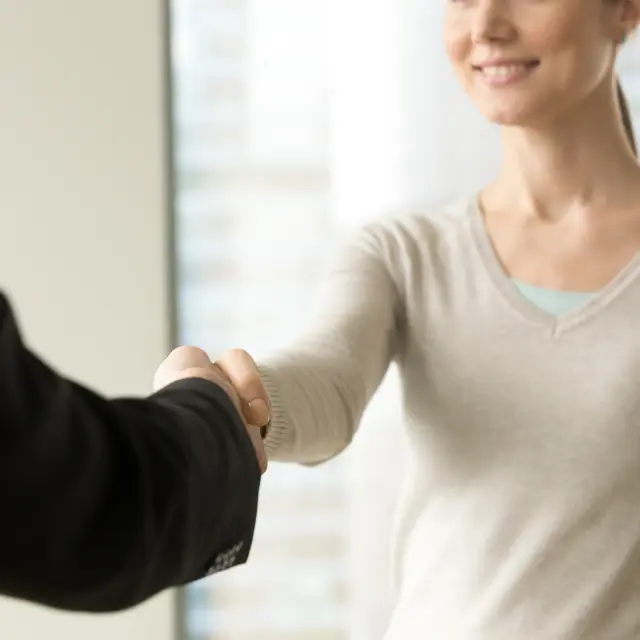 Une photo montrant une poignée de main entre deux personnes. Une femme souriante (visage partiellement visible, peau claire) porte un pull beige clair et tend la main; en face, un bras en veste noire serre sa main. L’arrière-plan de la photo est flou et clair, suggérant un intérieur lumineux.
