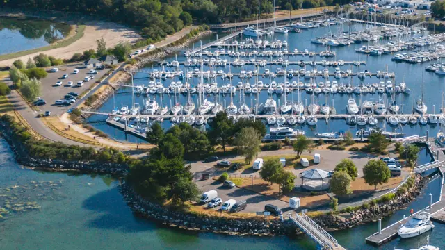 Photo d'un vue aérienne d’un port de plaisance. De nombreux voiliers sont amarrés dans des bassins. L’eau est bleu-vert au premier plan. Une jetée et des quais entourent le port, avec une zone de stationnement et quelques bâtiments. À l’arrière-plan, une bande de terre verdoyante avec des arbres et des champs, sous un ciel clair.