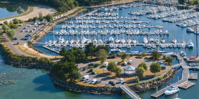 Photo d'un vue aérienne d’un port de plaisance. De nombreux voiliers sont amarrés dans des bassins. L’eau est bleu-vert au premier plan. Une jetée et des quais entourent le port, avec une zone de stationnement et quelques bâtiments. À l’arrière-plan, une bande de terre verdoyante avec des arbres et des champs, sous un ciel clair.