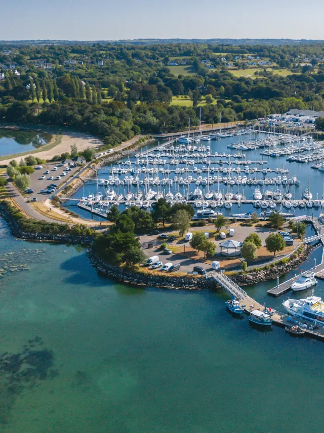 Photo d'un vue aérienne d’un port de plaisance. De nombreux voiliers sont amarrés dans des bassins. L’eau est bleu-vert au premier plan. Une jetée et des quais entourent le port, avec une zone de stationnement et quelques bâtiments. À l’arrière-plan, une bande de terre verdoyante avec des arbres et des champs, sous un ciel clair.