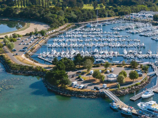 Luchtfoto van een jachthaven. Talloze zeilboten liggen aangemeerd in bassins. Het water is blauwgroen op de voorgrond. Een steiger en kades omringen de haven, met een parkeerplaats en een paar gebouwen. Op de achtergrond een groene strook land met bomen en akkers, onder een heldere hemel.