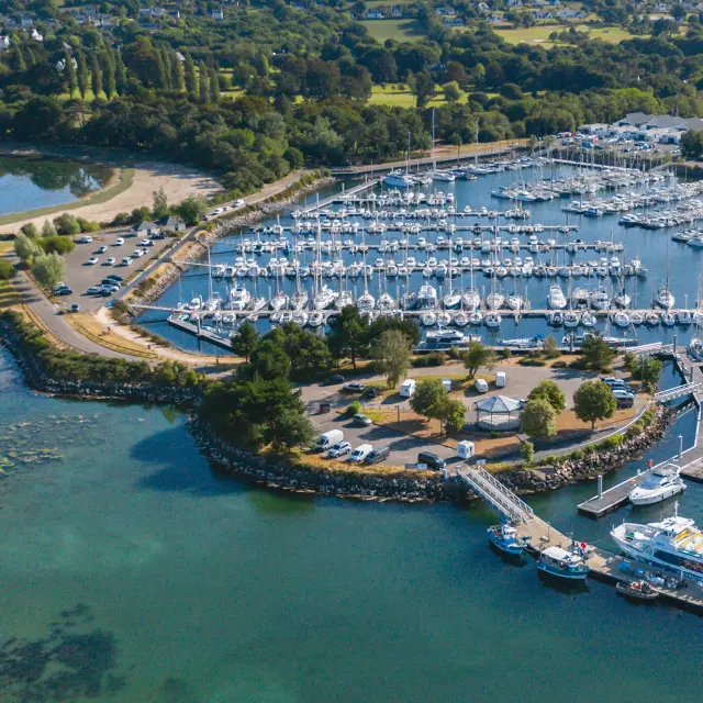Luchtfoto van een jachthaven. Talloze zeilboten liggen aangemeerd in bassins. Het water is blauwgroen op de voorgrond. Een steiger en kades omringen de haven, met een parkeerplaats en een paar gebouwen. Op de achtergrond een groene strook land met bomen en akkers, onder een heldere hemel.