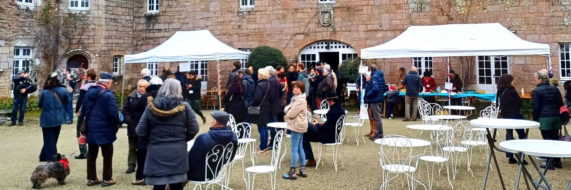 Une photo montrant un marché en extérieur devant un grand bâtiment en pierre de type manoir/château. La façade comporte plusieurs étages, de nombreuses fenêtres, et un toit sombre avec lucarnes. Au premier plan, plusieurs tentes blanches et des groupes de personnes en manteaux d’hiver. Des tables et chaises blanches sont disposées sur une cour en gravier.