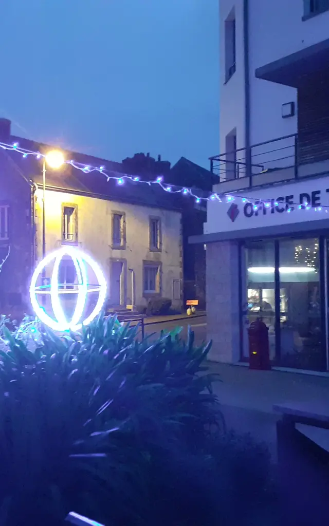 La photo montre une scène de nuit devant un bâtiment portant l’enseigne “OFFICE DE TOURISME”. Des décorations lumineuses de Noël bleues et blanches sont visibles: guirlandes et trois formes sphériques éclairées (type boules lumineuses) au premier plan, avec de la végétation sombre en bas de l’image. À droite, près de l’entrée, un panneau illustré est posé devant le bâtiment. Des lumières sont visibles à l’intérieur derrière les vitrines.