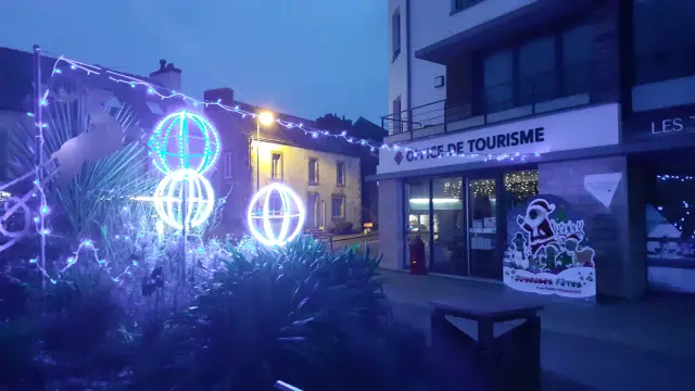 De foto toont een nachtscène voor een gebouw met het bord OFFICE DE TOURISME. Blauwe en witte kerstversieringen zijn zichtbaar: slingers en drie verlichte bolvormen (kerstballen) op de voorgrond, met donkere begroeiing onderaan het beeld. Rechts, bij de ingang, staat een geïllustreerd bord voor het gebouw. Binnen zijn lichten zichtbaar achter de etalages.