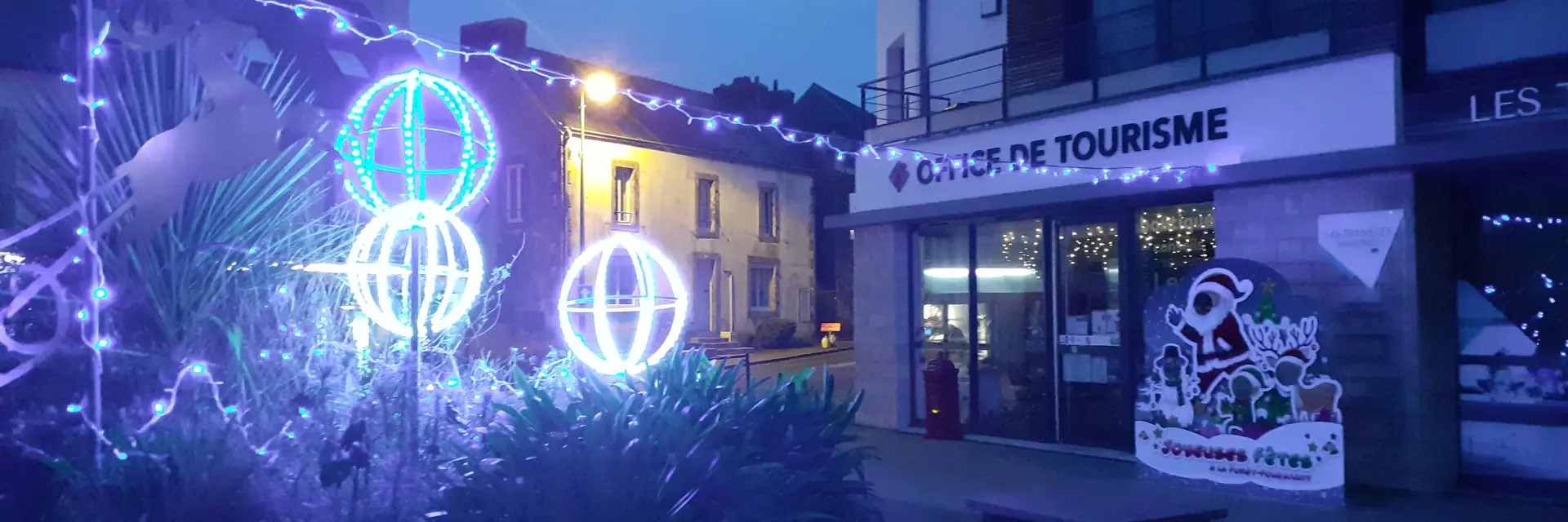 La photo montre une scène de nuit devant un bâtiment portant l’enseigne “OFFICE DE TOURISME”. Des décorations lumineuses de Noël bleues et blanches sont visibles: guirlandes et trois formes sphériques éclairées (type boules lumineuses) au premier plan, avec de la végétation sombre en bas de l’image. À droite, près de l’entrée, un panneau illustré est posé devant le bâtiment. Des lumières sont visibles à l’intérieur derrière les vitrines.