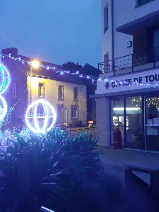 La photo montre une scène de nuit devant un bâtiment portant l’enseigne “OFFICE DE TOURISME”. Des décorations lumineuses de Noël bleues et blanches sont visibles: guirlandes et trois formes sphériques éclairées (type boules lumineuses) au premier plan, avec de la végétation sombre en bas de l’image. À droite, près de l’entrée, un panneau illustré est posé devant le bâtiment. Des lumières sont visibles à l’intérieur derrière les vitrines.