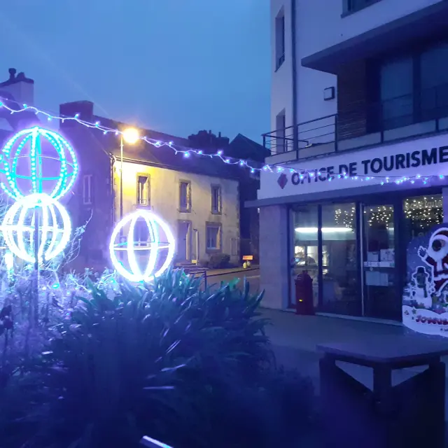 De foto toont een nachtscène voor een gebouw met het bord OFFICE DE TOURISME. Blauwe en witte kerstversieringen zijn zichtbaar: slingers en drie verlichte bolvormen (kerstballen) op de voorgrond, met donkere begroeiing onderaan het beeld. Rechts, bij de ingang, staat een geïllustreerd bord voor het gebouw. Binnen zijn lichten zichtbaar achter de etalages.