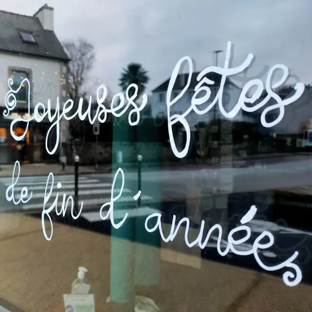 La photo montre une vitrine avec l’inscription en écriture blanche “Joyeuses fêtes de fin d’année” ; en arrière-plan flou, des bâtiments de rue et des décorations lumineuses.