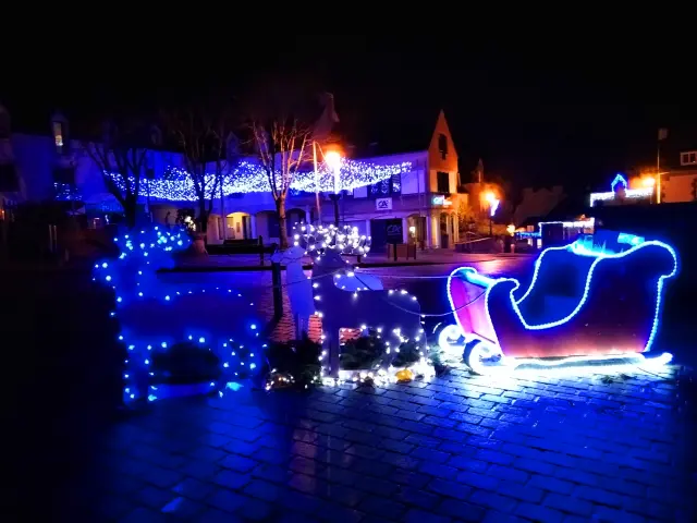 Une photo de décorations de Noël de nuit. La photo montre une place avec des illuminations bleues et blanches, notamment une silhouette lumineuse de renne à gauche et un traîneau lumineux à droite, avec des bâtiments en arrière-plan décorés de guirlandes.