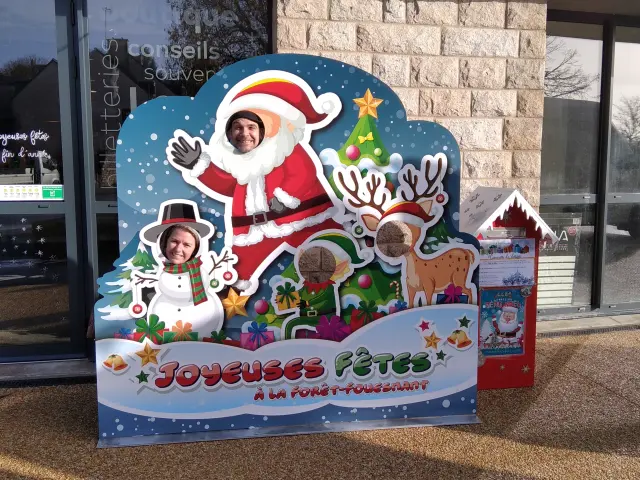 La photo montre une grande décoration de Noël “Joyeuses Fêtes” placée devant un bâtiment, avec un Père Noël, un renne, un bonhomme de neige et un sapin stylisés. En bas de la photo, un texte “Joyeuses Fêtes” est lisible.