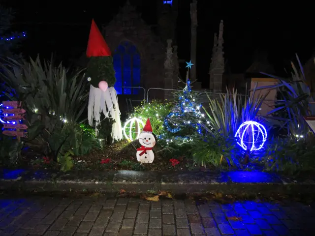 La photo montre une décoration de Noël de nuit dans un massif de plantes: un petit bonhomme de neige éclairé au premier plan, un sapin lumineux avec une étoile au sommet au centre, des guirlandes et points lumineux blancs autour des plantes, et deux grandes décorations lumineuses en forme de sphères (une blanche à gauche du sapin, une bleue à droite). À gauche, une grande silhouette décorative type personnage/gnome avec un bonnet rouge pointu et une longue barbe blanche. Le sol au bas de la photo est un pavage mouillé reflétant des lumières bleues.