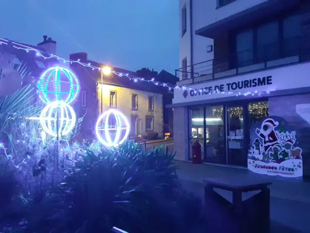 La photo montre une scène de nuit devant un bâtiment portant l’enseigne “OFFICE DE TOURISME”. Des décorations lumineuses de Noël bleues et blanches sont visibles: guirlandes et trois formes sphériques éclairées (type boules lumineuses) au premier plan, avec de la végétation sombre en bas de l’image. À droite, près de l’entrée, un panneau illustré est posé devant le bâtiment. Des lumières sont visibles à l’intérieur derrière les vitrines.