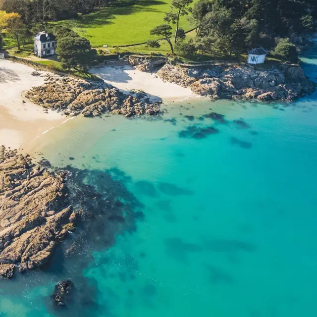 Ein Foto einer Bucht, das die Luftaufnahme einer Küste mit kleinen hellen Sandstränden, Felsen, türkisfarbenem Wasser und einem Waldgebiet mit einer großen grünen Wiese zeigt; ein helles Gebäude steht in der Nähe des Ufers.