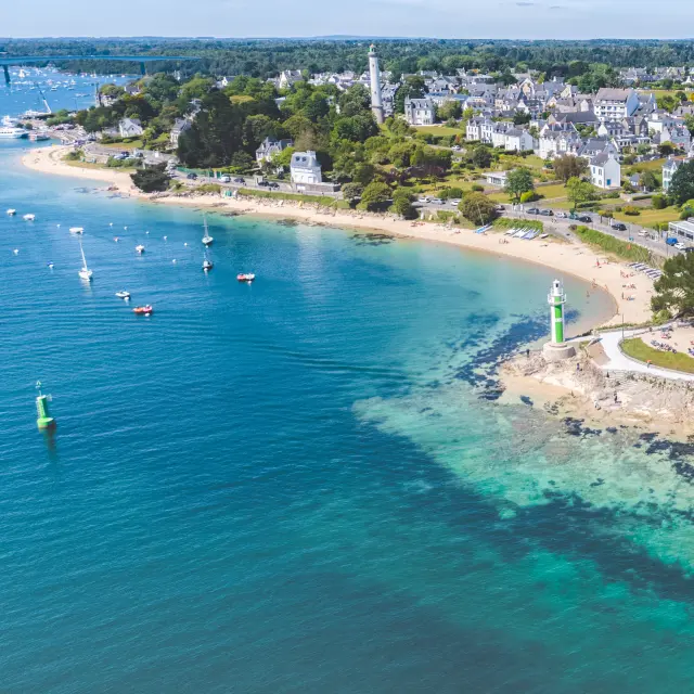 Ein Luftbild einer Küste mit einer Bucht mit blauem/türkisfarbenem Wasser, ankernden Booten, einem gebogenen Strand, einem kleinen Leuchtturm/grünem Feuer auf einem Steg und dann einer Küstenstadt mit weißen Häusern und Bäumen.