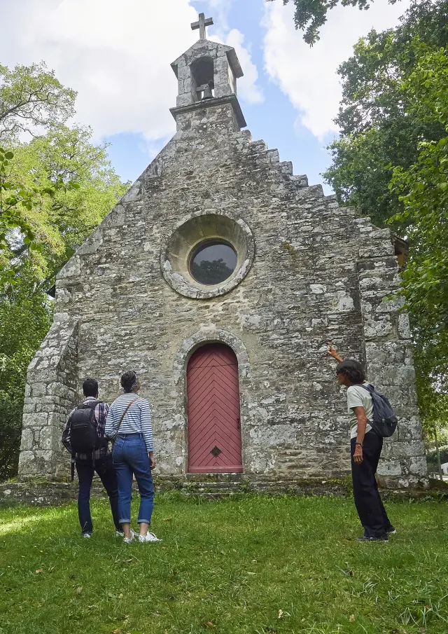 Ein Foto einer kleinen Steinkapelle von vorne, mit einem Glockenturm, auf dem ein Kreuz steht. Die Fassade hat ein rundes Fenster über einer roten Tür. Grüne Bäume rahmen die Kapelle ein und im Vordergrund befindet sich eine Rasenfläche. Drei Personen stehen vor der Tür.