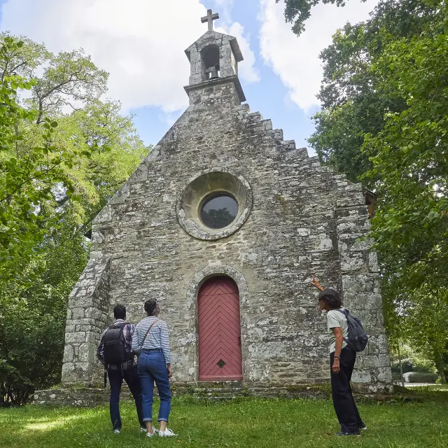 Une photo d'une petite chapelle en pierre vue de face, avec un clocher surmonté d’une croix. La façade a une fenêtre ronde au-dessus d’une porte rouge. Des arbres verts encadrent la chapelle et une pelouse est au premier plan. Trois personnes se tiennent devant la porte.