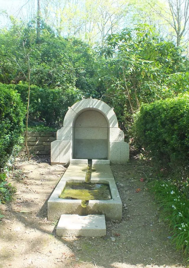 Das Foto zeigt einen Steinbrunnen am Ende eines kleinen Weges, der von grünen Hecken umgeben ist. Der Brunnen hat ein bogenförmiges Oberteil, einen Ausguss in der Mitte und unten ein rechteckiges Becken mit Wasser. Dahinter sind dichte Vegetation und Bäume zu sehen.