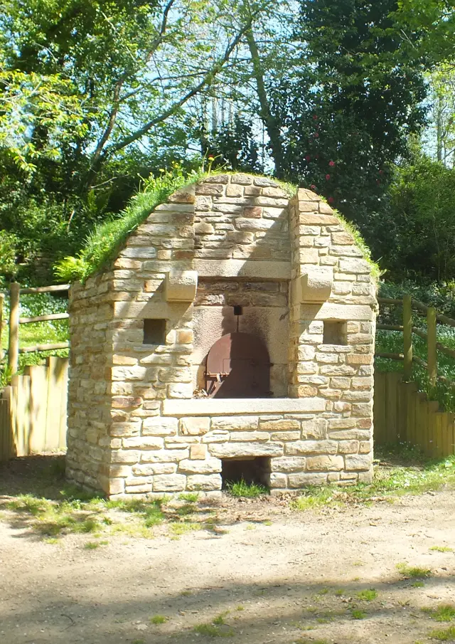 Ein Foto von einem Steinofen (Brotbackofen) in einem Unterholz. Der Ofen befindet sich links im Vordergrund und hat eine dunkle Öffnung. Ein unbefestigter Weg führt nach rechts zu einem zweiten kleinen Ofen, der im Hintergrund zu sehen ist. Laubbäume und Vegetation umgeben die Szene, die von der Sonne beleuchtet wird.