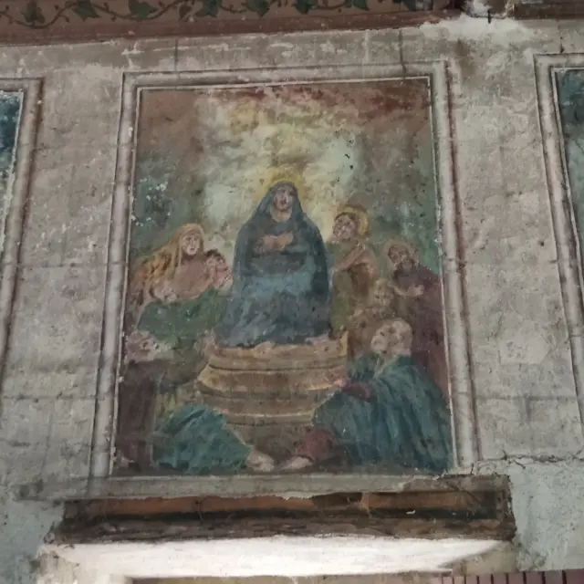 Foto eines alten und teilweise verfallenen Wandfreskos, das von bemalten Zierleisten eingerahmt wird. Die Szene zeigt mehrere Personen um eine zentrale Figur herum, die in gedämpften Farben (Grün, Blau, Ocker) dargestellt sind. Das Fresko befindet sich über einer Türöffnung, die am unteren Rand des Bildes zu sehen ist.
