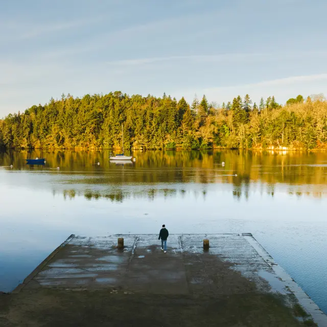 Une photo qui montre un plan d’eau très calme, avec une forêt de conifères au fond éclairée par une lumière dorée. Au premier plan, une cale en béton avance dans l’eau. Une personne seule, de petite taille dans l’image, se tient au bout de la cale, face au lac. La surface de l’eau reflète les arbres et le ciel. Le ciel est bleu clair, sans nuages marqués.