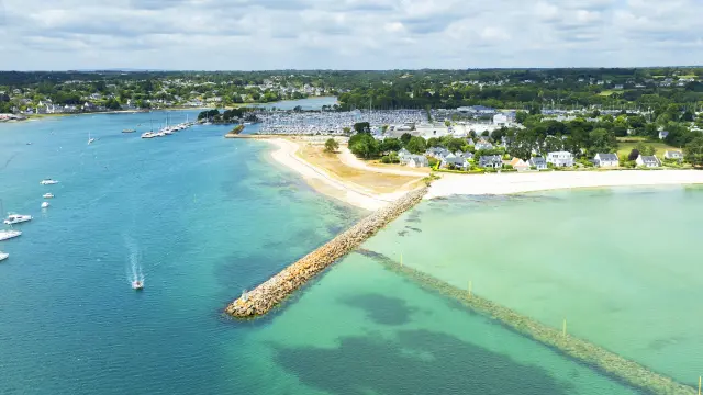 Photo aérienne d’un littoral qui montre une mer turquoise et bleu foncé, une longue digue en pierres qui avance dans l’eau, une plage claire en arc, et au fond une zone de port avec de nombreux bateaux/mâts, puis des maisons et de la végétation sous un ciel bleu avec des nuages.