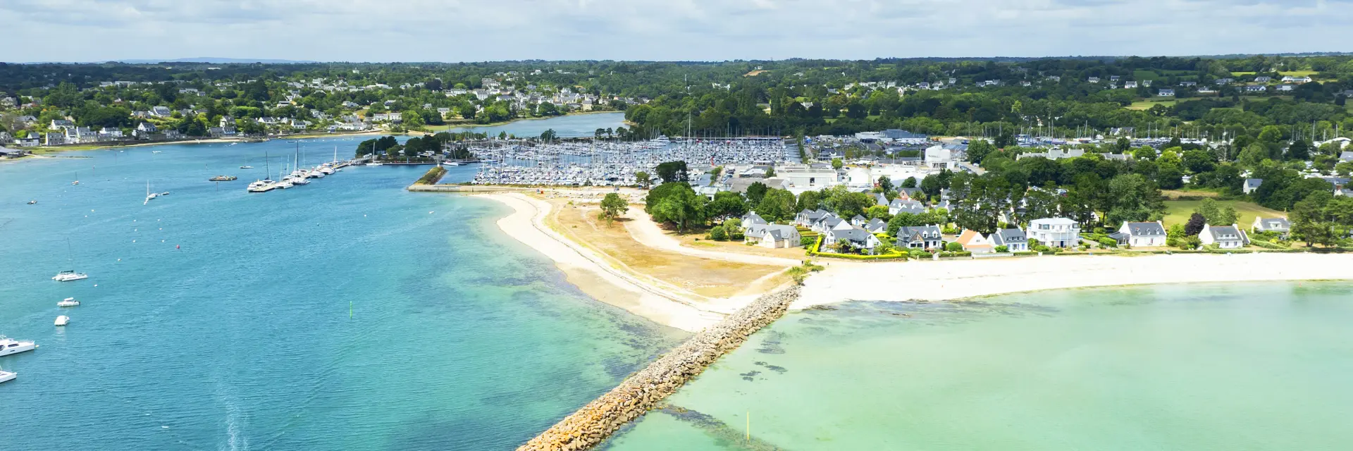 Photo aérienne d’un littoral qui montre une mer turquoise et bleu foncé, une longue digue en pierres qui avance dans l’eau, une plage claire en arc, et au fond une zone de port avec de nombreux bateaux/mâts, puis des maisons et de la végétation sous un ciel bleu avec des nuages.