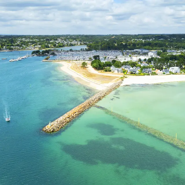 Photo aérienne d’un littoral qui montre une mer turquoise et bleu foncé, une longue digue en pierres qui avance dans l’eau, une plage claire en arc, et au fond une zone de port avec de nombreux bateaux/mâts, puis des maisons et de la végétation sous un ciel bleu avec des nuages.