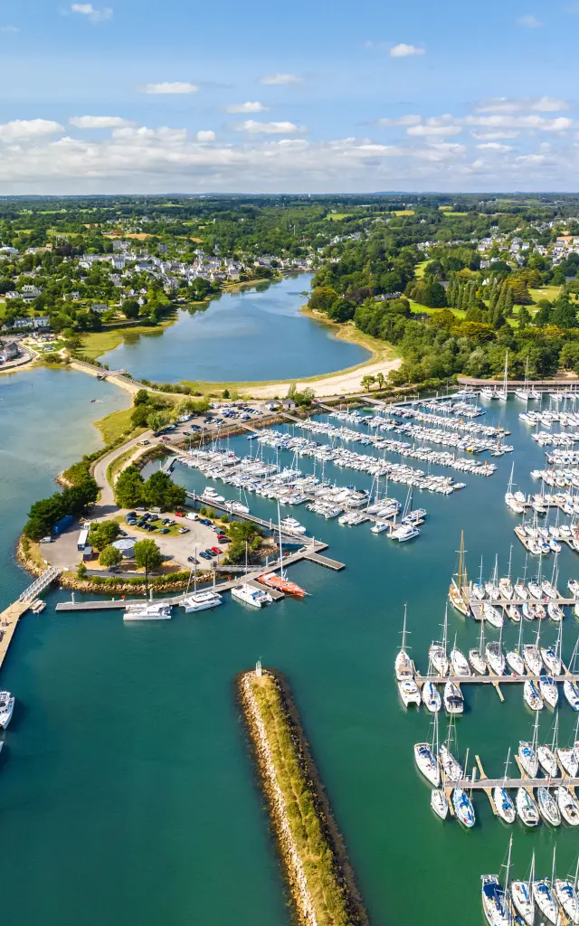 Photo aérienne d’un port de plaisance qui montre une baie avec eau bleu-vert, une digue, des pontons et de très nombreux voiliers alignés; au fond, des zones vertes et des habitations sous un ciel bleu avec nuages.