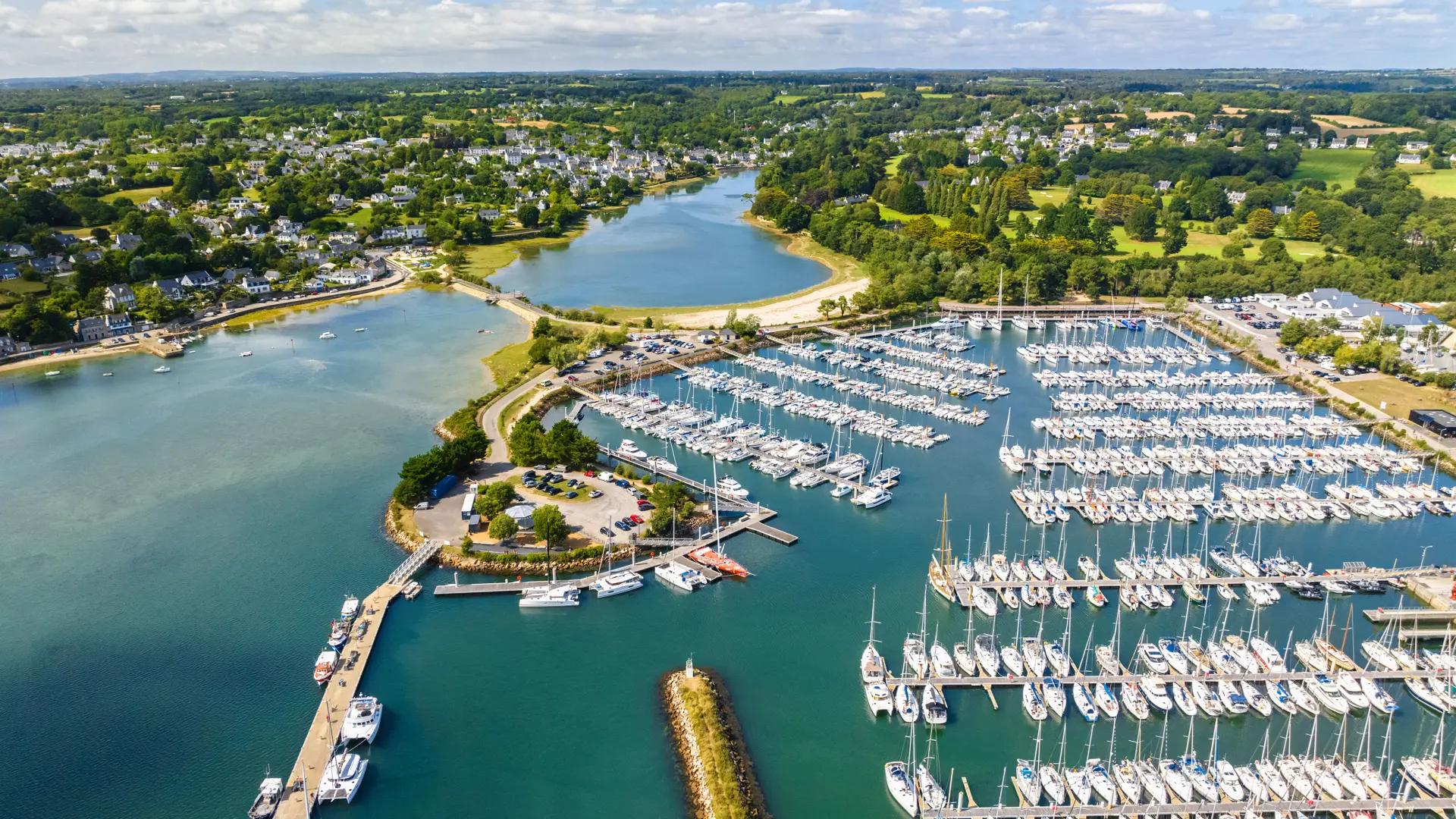 Photo aérienne d’un port de plaisance qui montre une baie avec eau bleu-vert, une digue, des pontons et de très nombreux voiliers alignés; au fond, des zones vertes et des habitations sous un ciel bleu avec nuages.
