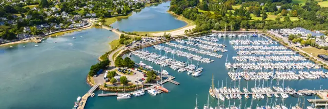 Photo aérienne d’un port de plaisance qui montre une baie avec eau bleu-vert, une digue, des pontons et de très nombreux voiliers alignés; au fond, des zones vertes et des habitations sous un ciel bleu avec nuages.
