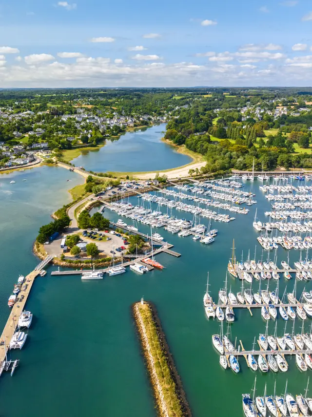 Photo aérienne d’un port de plaisance qui montre une baie avec eau bleu-vert, une digue, des pontons et de très nombreux voiliers alignés; au fond, des zones vertes et des habitations sous un ciel bleu avec nuages.