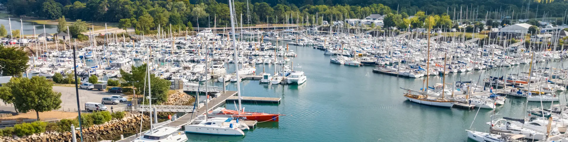 Photo d’un port de plaisance rempli de voiliers et bateaux amarrés, une eau vert-turquoise, une digue en pierres, et un voilier au premier plan; au fond, une zone boisée et un ciel avec de gros nuages.