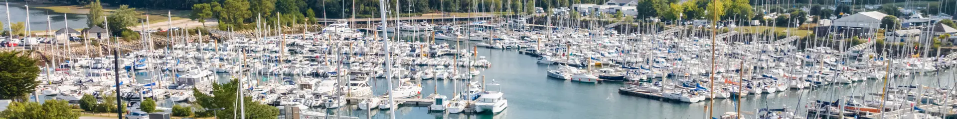 Photo d’un port de plaisance rempli de voiliers et bateaux amarrés, une eau vert-turquoise, une digue en pierres, et un voilier au premier plan; au fond, une zone boisée et un ciel avec de gros nuages.
