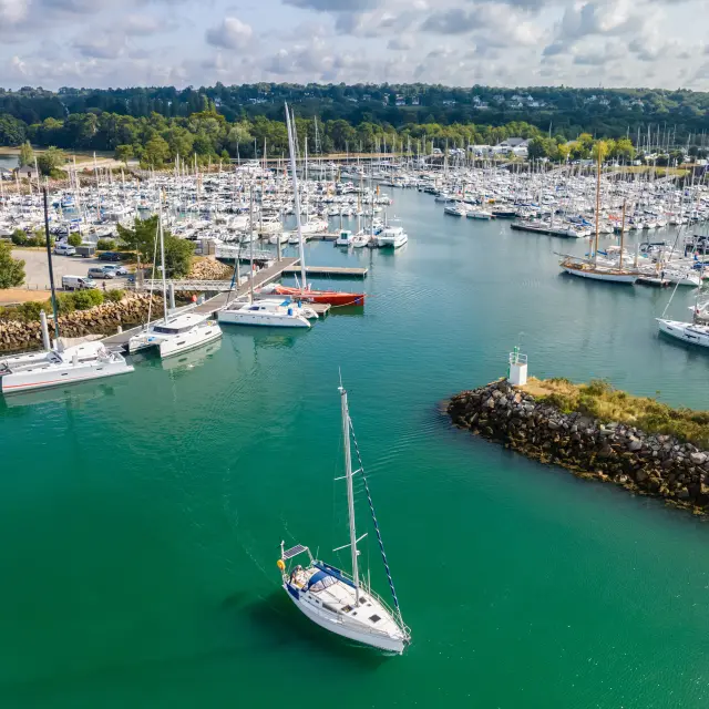 Photo d’un port de plaisance rempli de voiliers et bateaux amarrés, une eau vert-turquoise, une digue en pierres, et un voilier au premier plan; au fond, une zone boisée et un ciel avec de gros nuages.